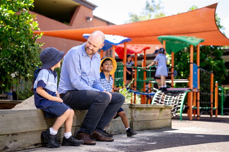 A male teacher sitting with two students in the playground